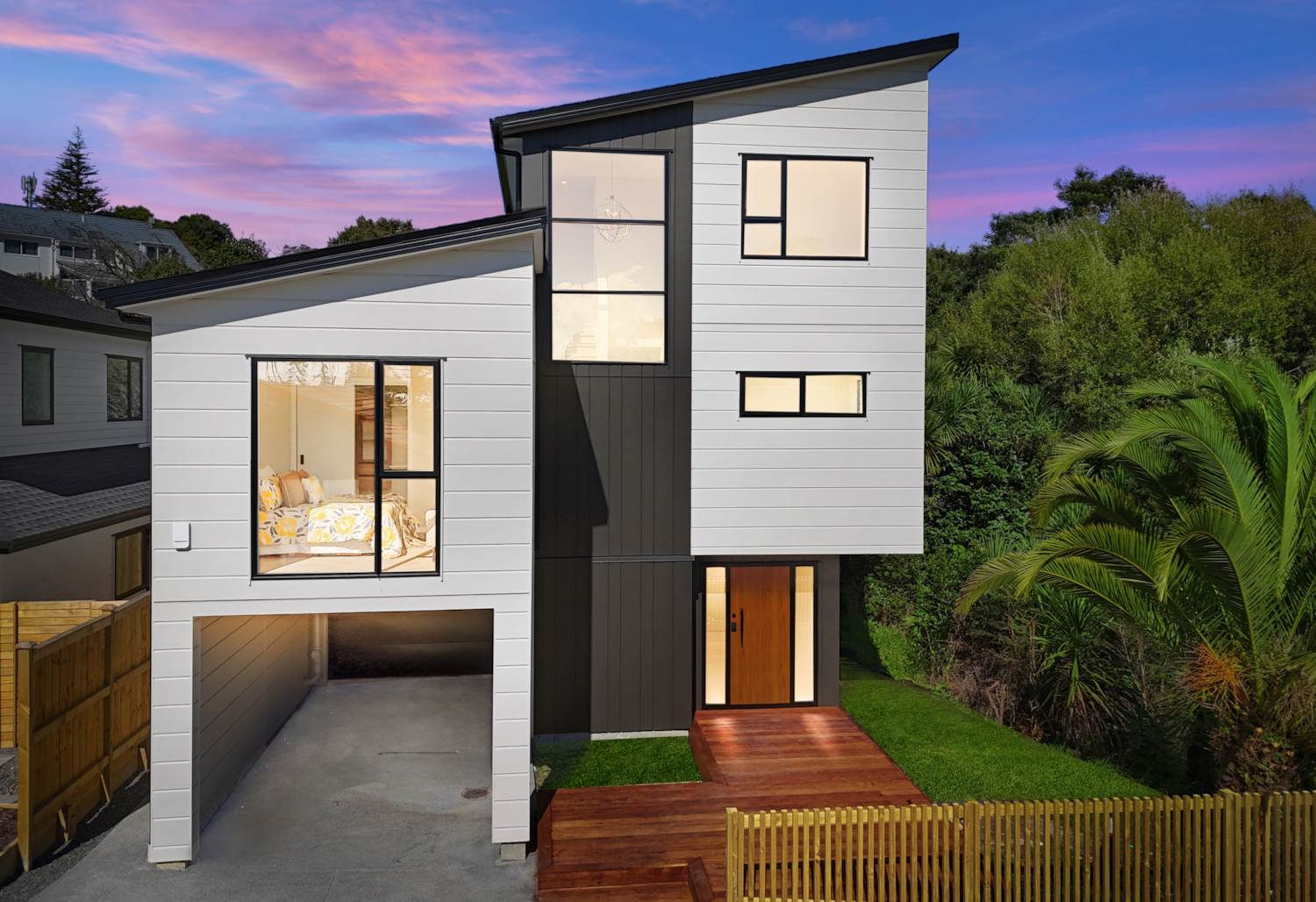 Three-level house with dark vertical cladding and a timber entry deck at dusk, Browns Bay