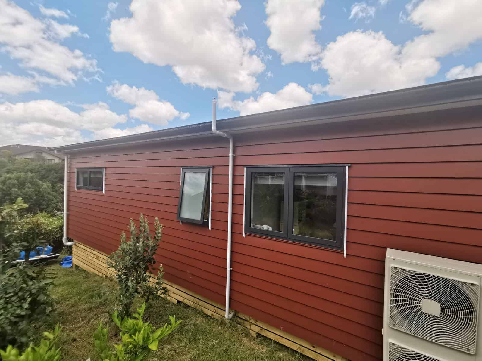 Long side elevation in red weatherboard with a heat pump unit at ground level, Botany Downs