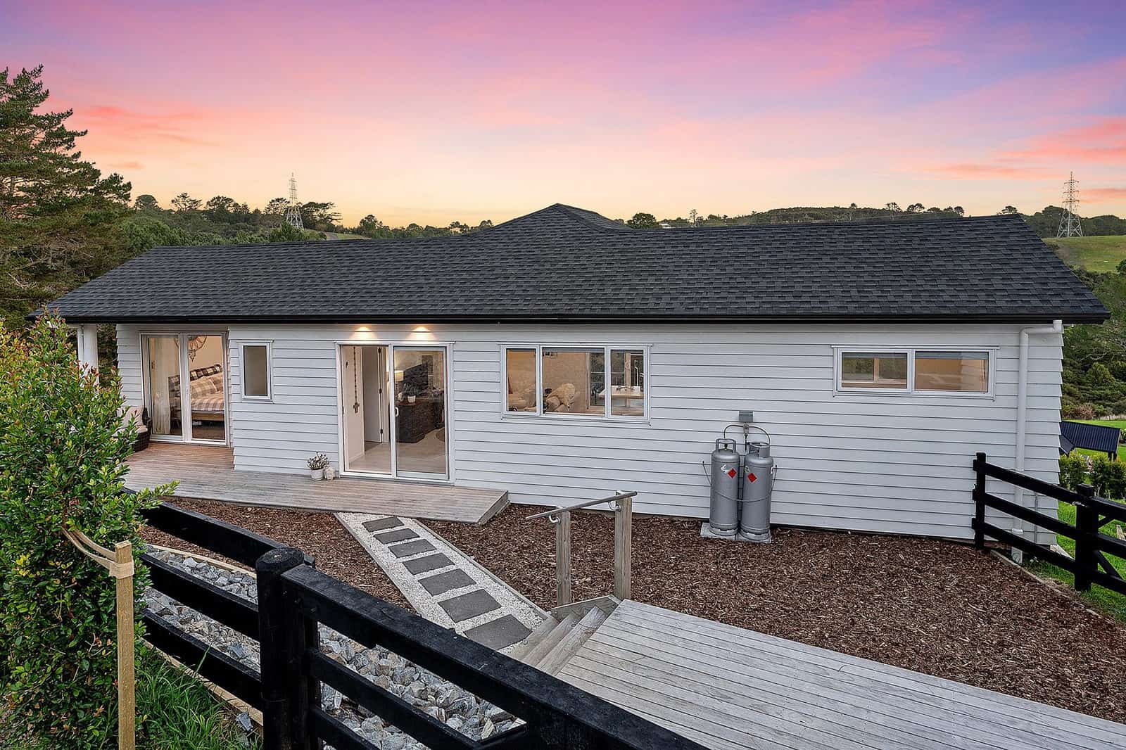 Single-level minor dwelling with a dark roof and deck seen across a post-and-rail fence at dusk, Coatesville