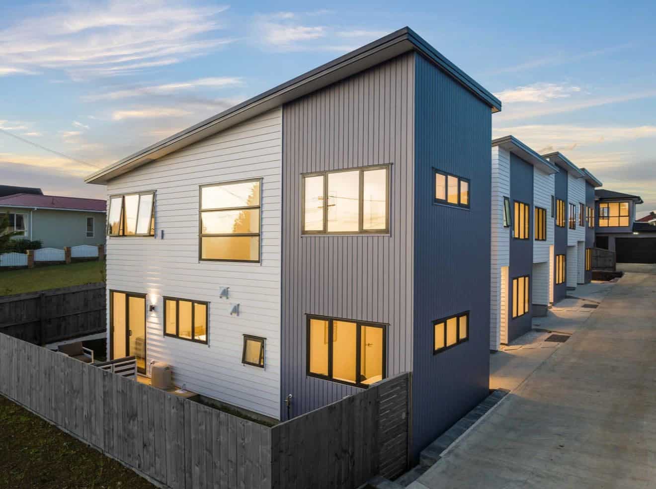 Row of new two-storey houses lit at dusk behind a timber fence, Lynfield
