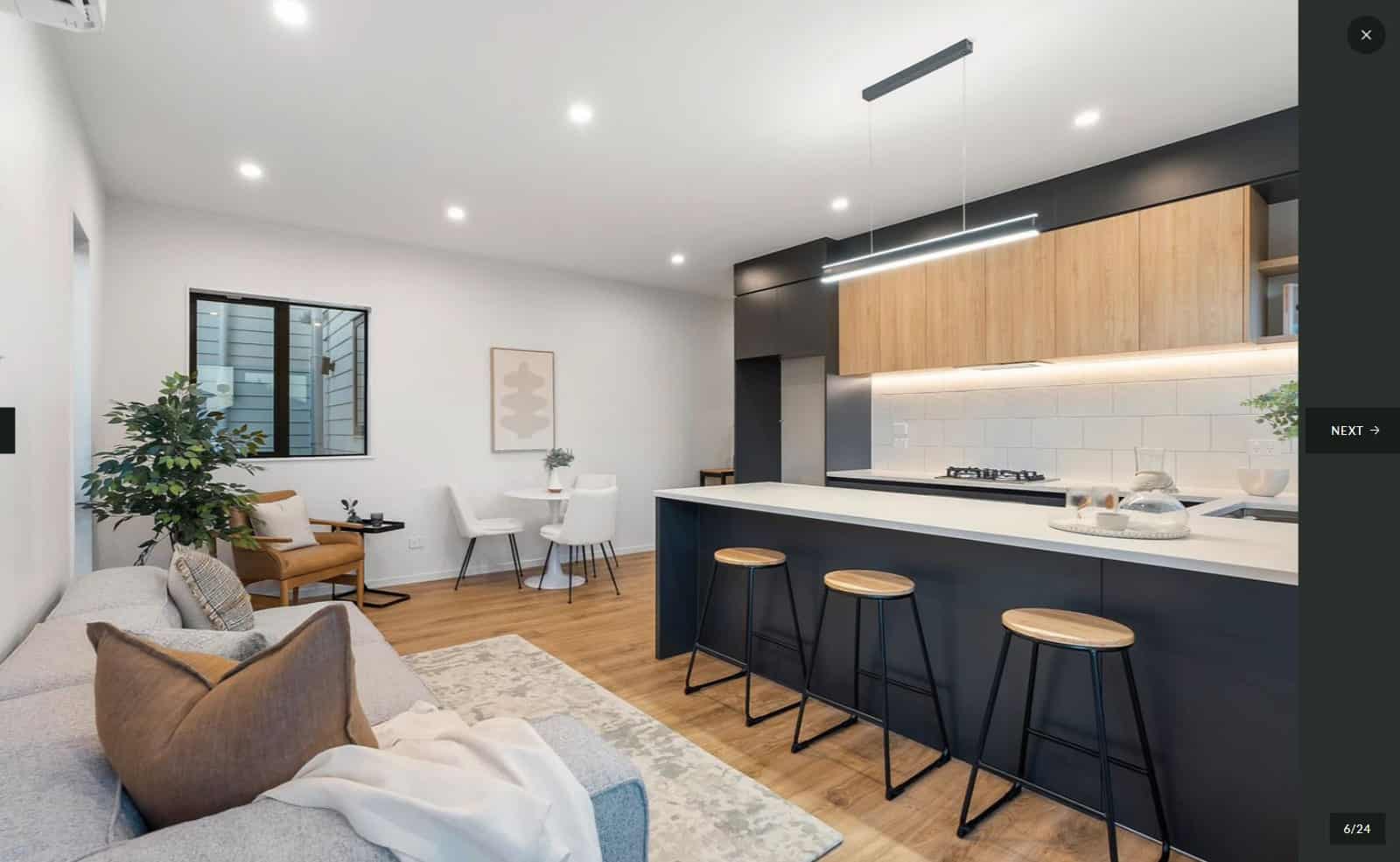 Open-plan kitchen and living with a dark island, timber overhead cabinetry and bar stools, Lynfield