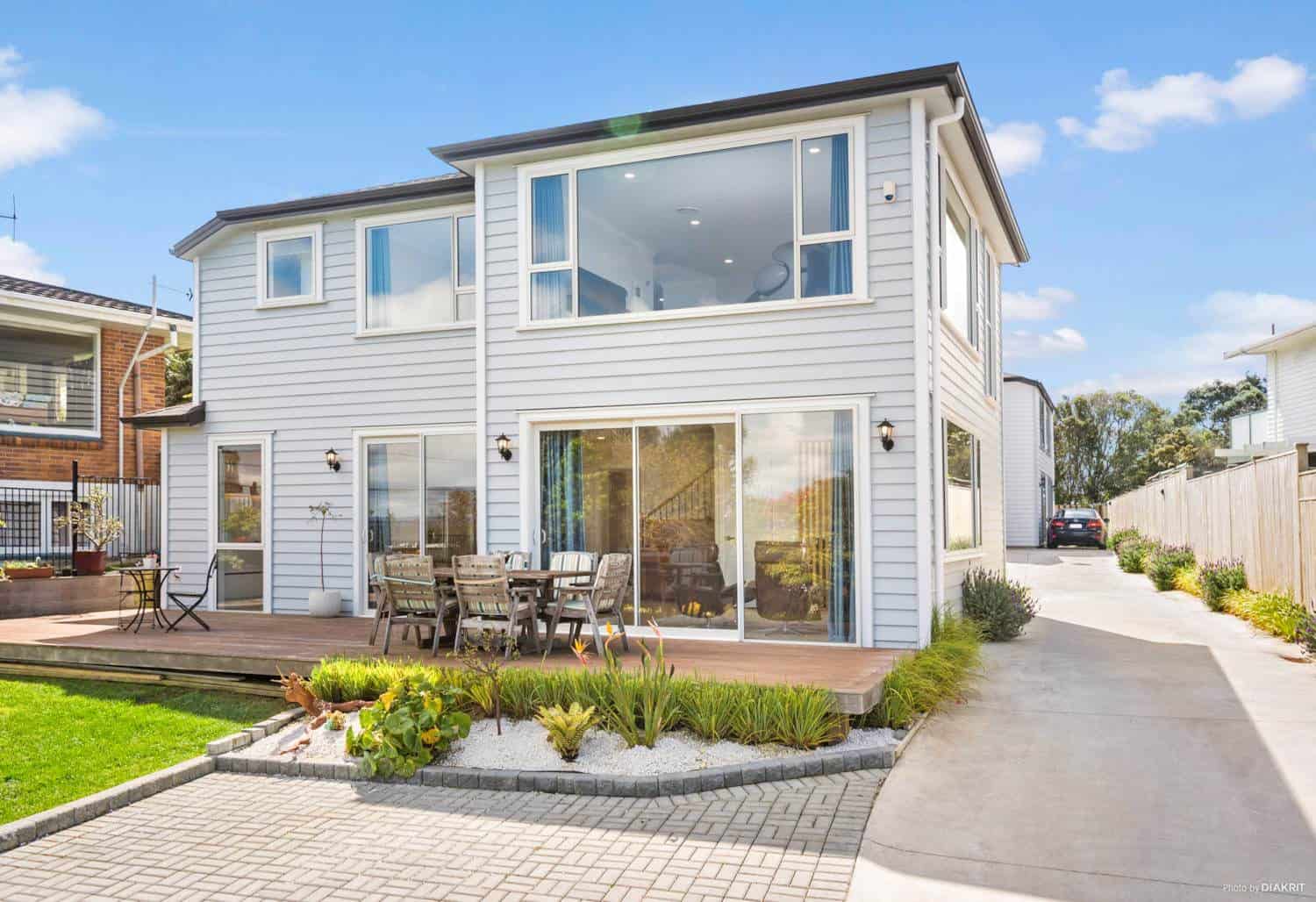 Two-storey weatherboard house with a deck, paved drive and planted garden, Mairangi Bay