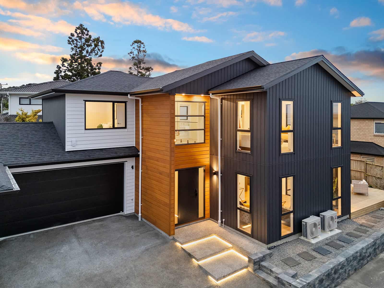 Two-storey home in dark cladding with a timber feature panel, lit at dusk, New Lynn