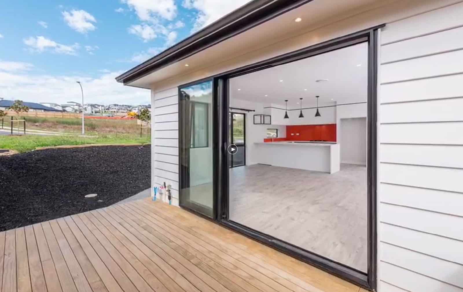 Sliding doors open from the deck into a kitchen with red cabinetry, Orewa