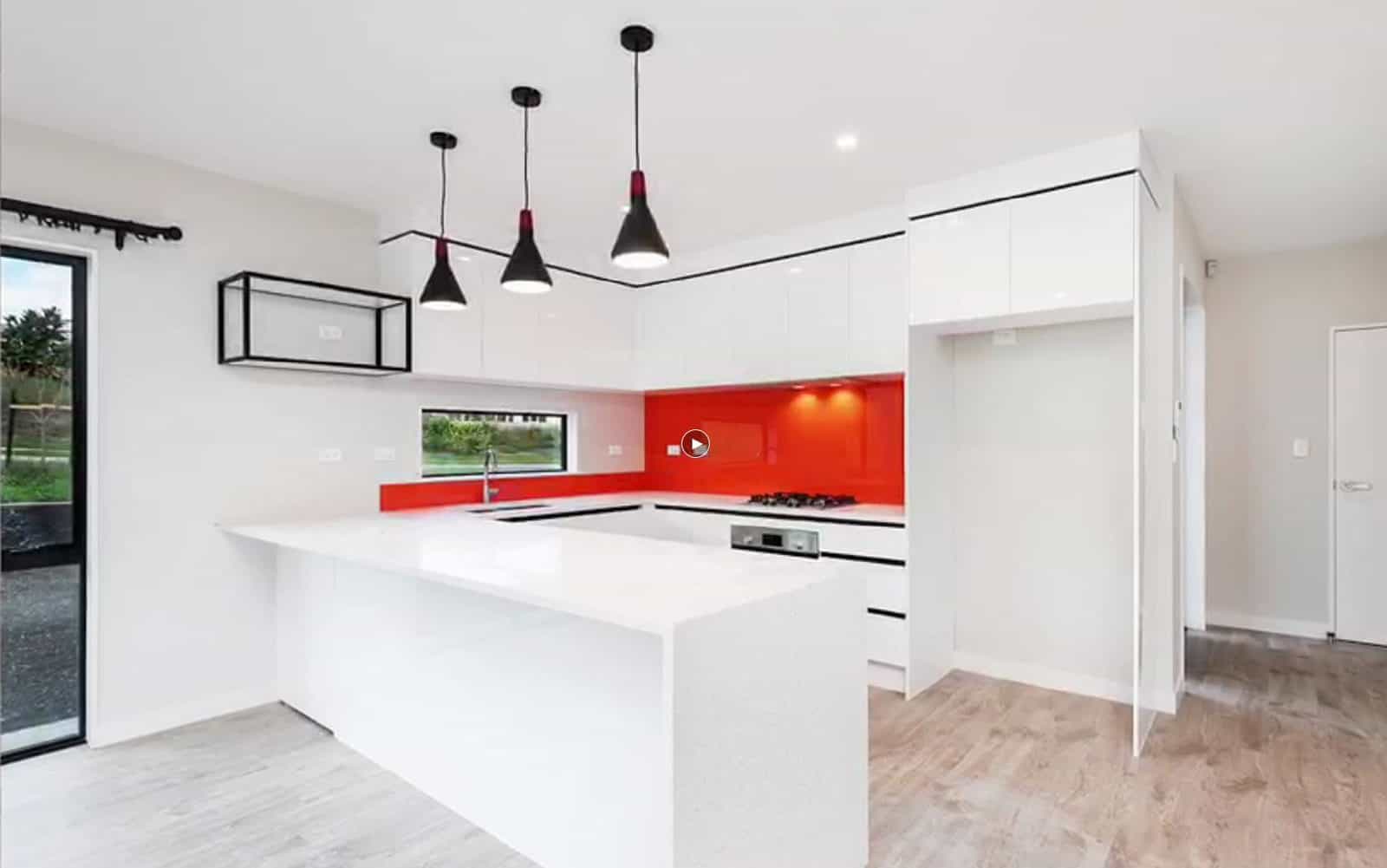 Kitchen with a white island, red glass splashback and pendant lights, Orewa
