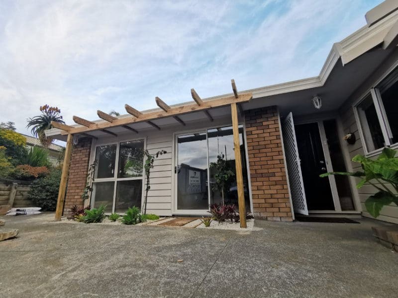 Former garage front rebuilt with weatherboards, a glazed window and a sliding door under a new timber pergola, beside the brick house entry