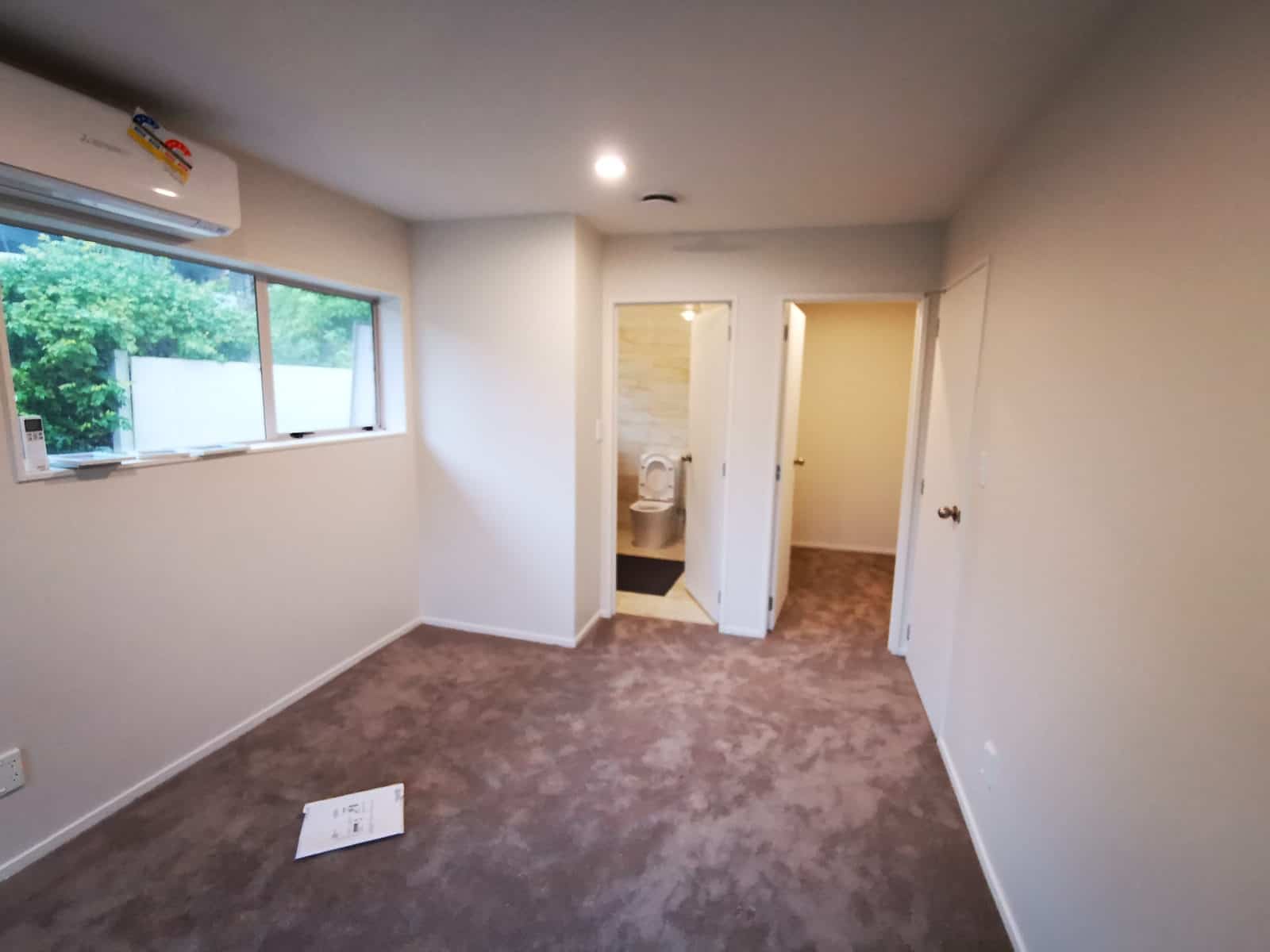 Carpeted room inside the converted garage with a wall heat pump, a window and doors through to a toilet and a hallway